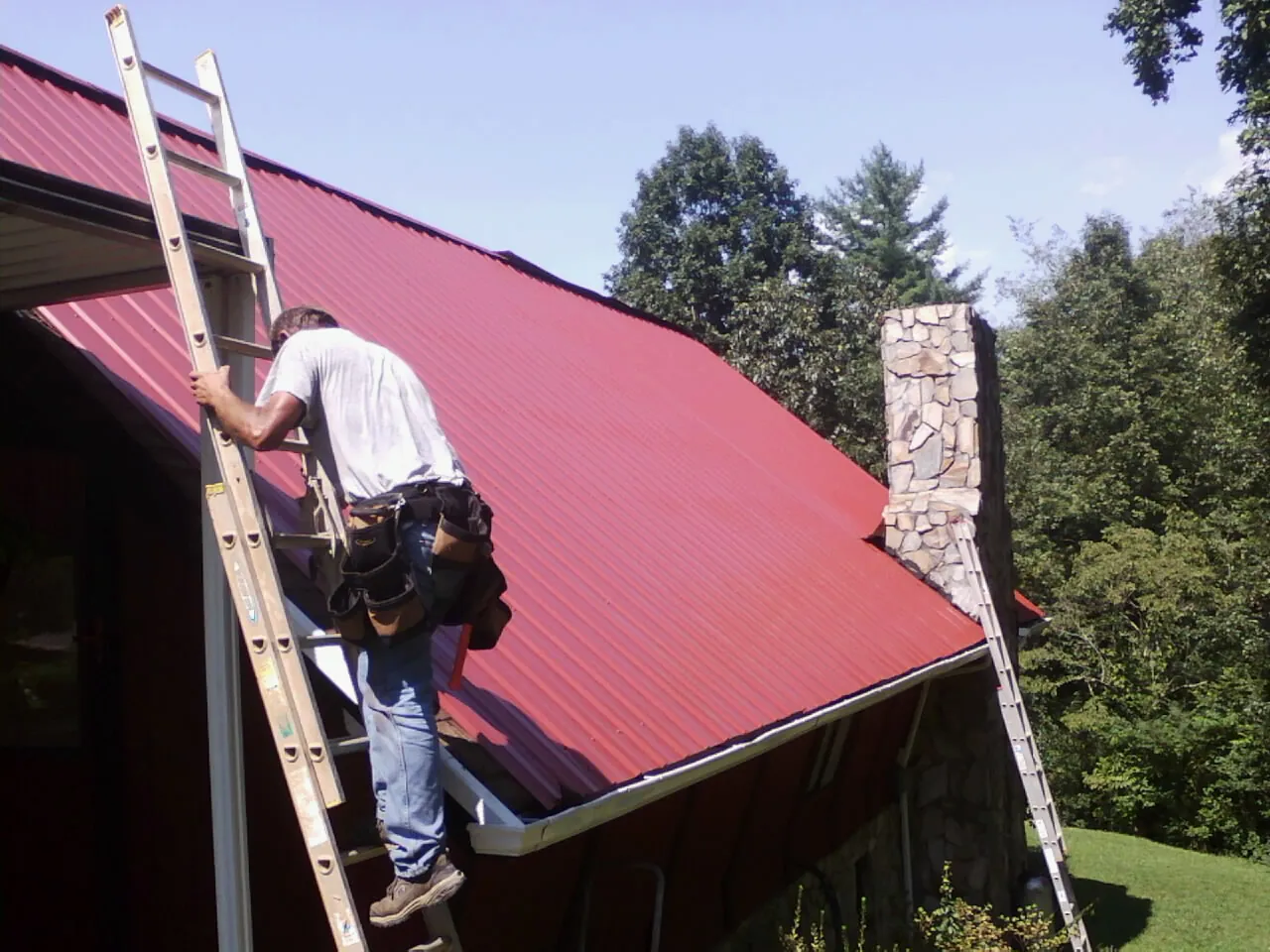 Roofer on a ladder beside a red metal roof and stone chimney — Soffit & Fascia Repair by Roofing Brothers in Tahoma